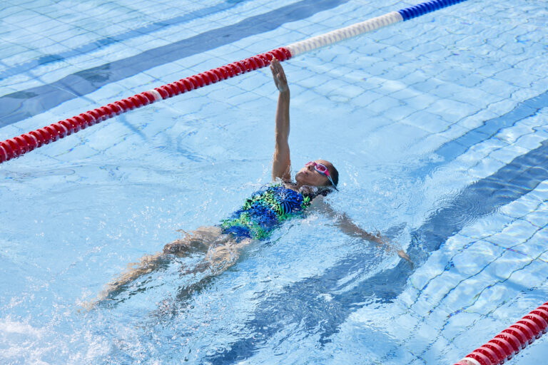 Photo looking downwards upon a young girl swimming backstroke in a swimming pool, with one arm oustretched above water.